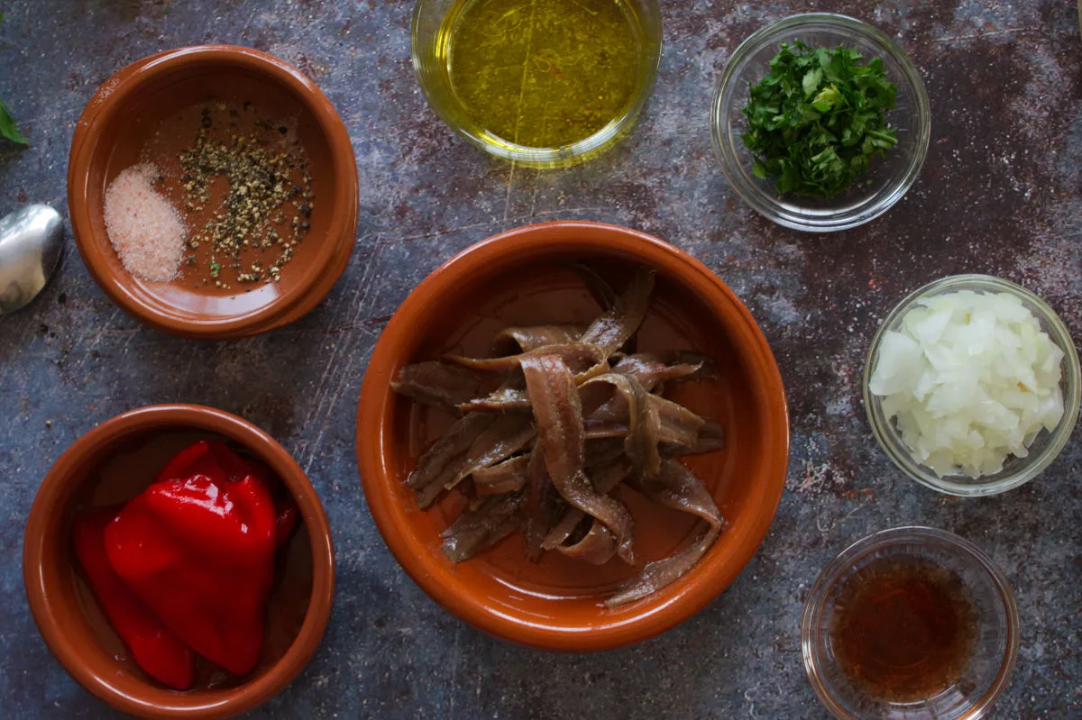 A small bowl of anchovies sits beside some roasted piquillo pepper, olive oil, fresh parsley, and onion. 