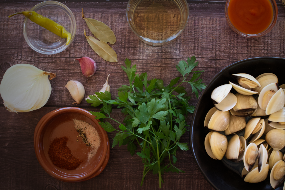 A bowl of fresh clams sits beside spices, fresh parsley, and some wine and tomato paste. 