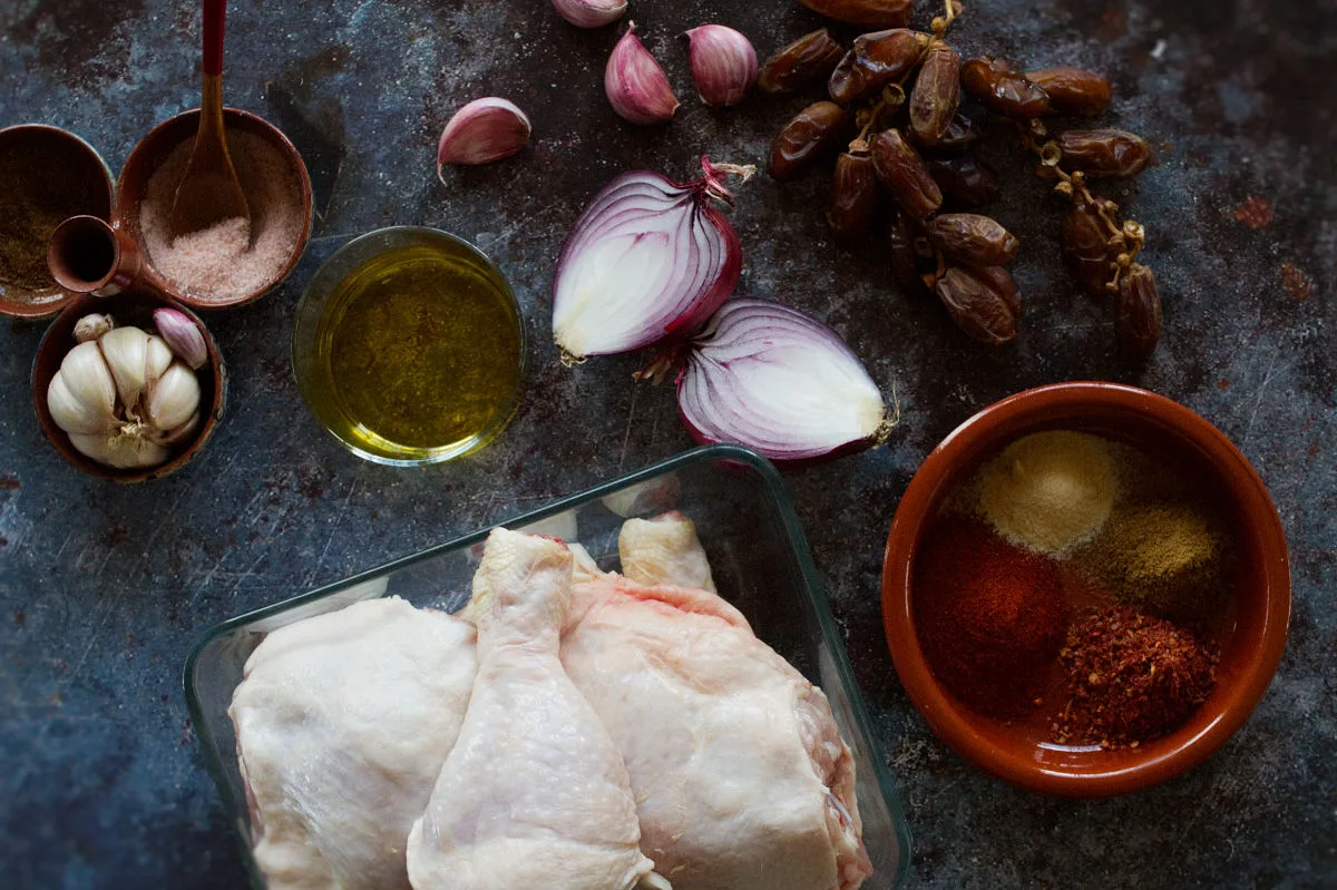 chicken pieces, onion, dates, herbs and spices sit on a kitchen counter. 