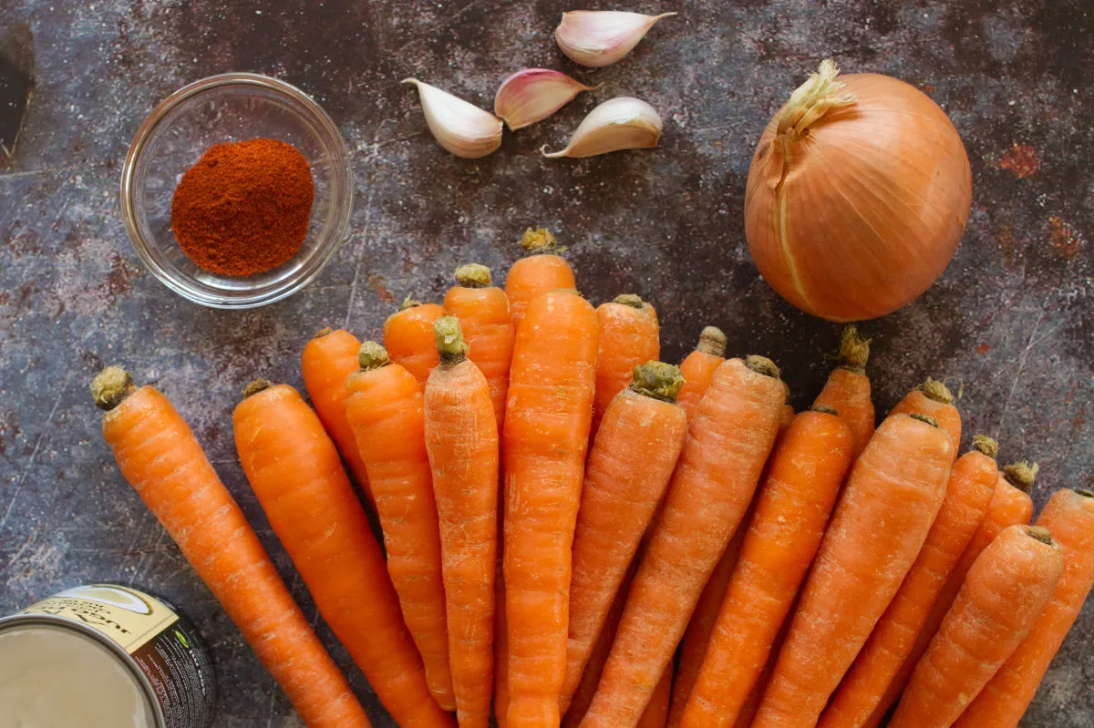 A pile of carrots sits beside an onion, some garlic cloves, and some smoked paprika.