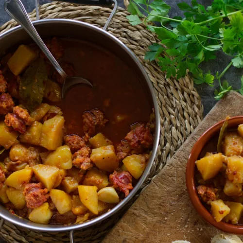 A large pot of Rioja-style potatoes with chorizo sits beside a small bowl serving.