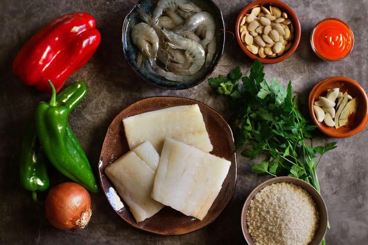 a plate of cod fish sits beside vegetables, shrimp, almonds, fresh herbs, and spices. 