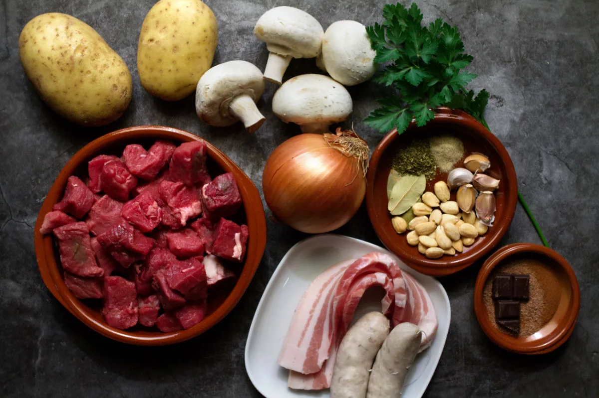 Chuck steak, butifarra sausage, pancetta and vegetables sit on a counter top.