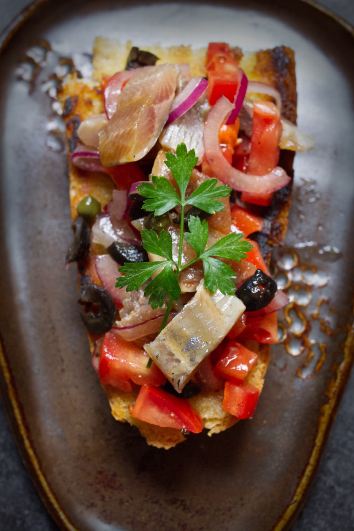 A slice of toast topped with tomato and sardiens with capers and olives