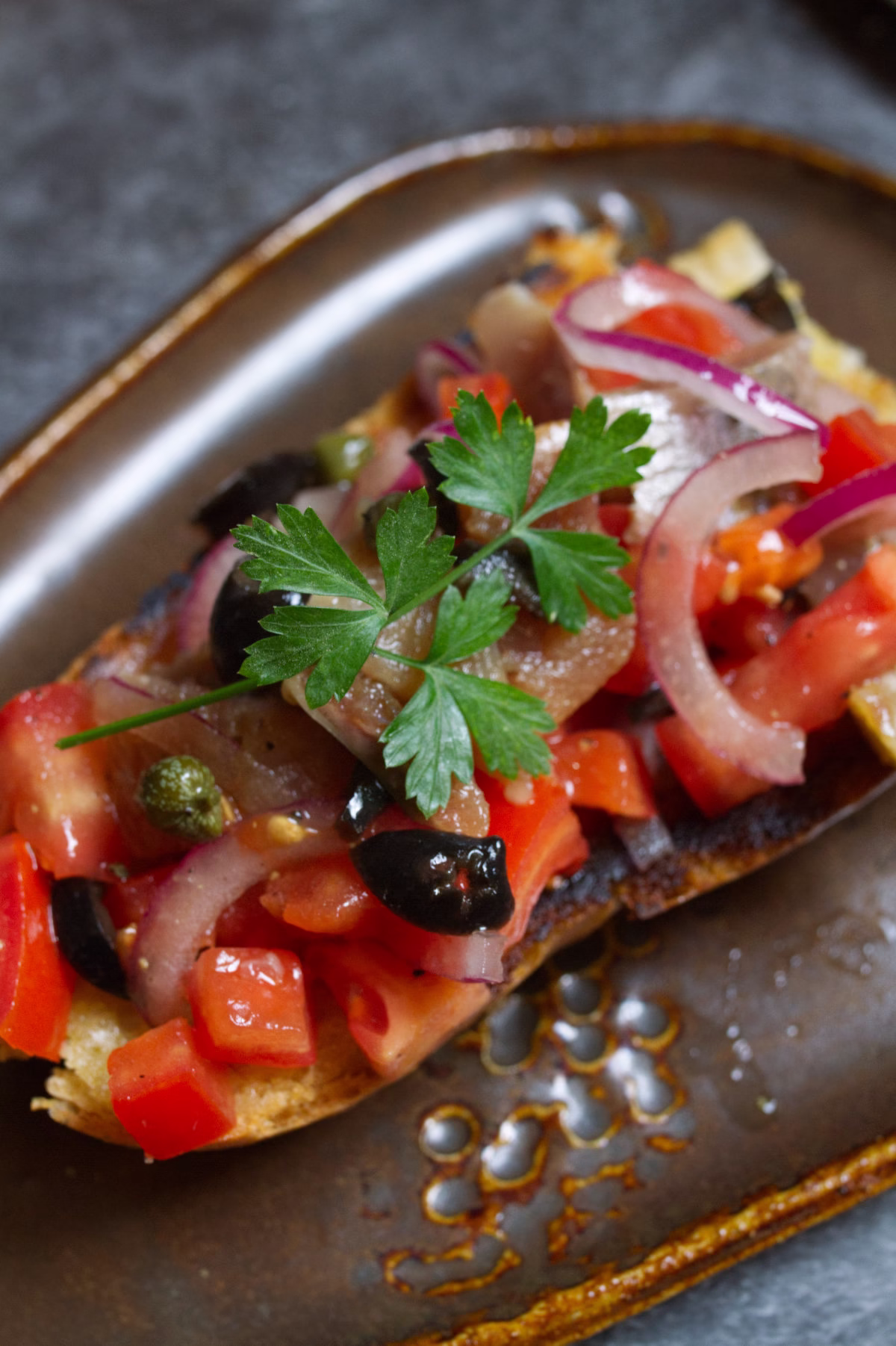 A slice of toast topped with tomato and sardiens with capers and olives