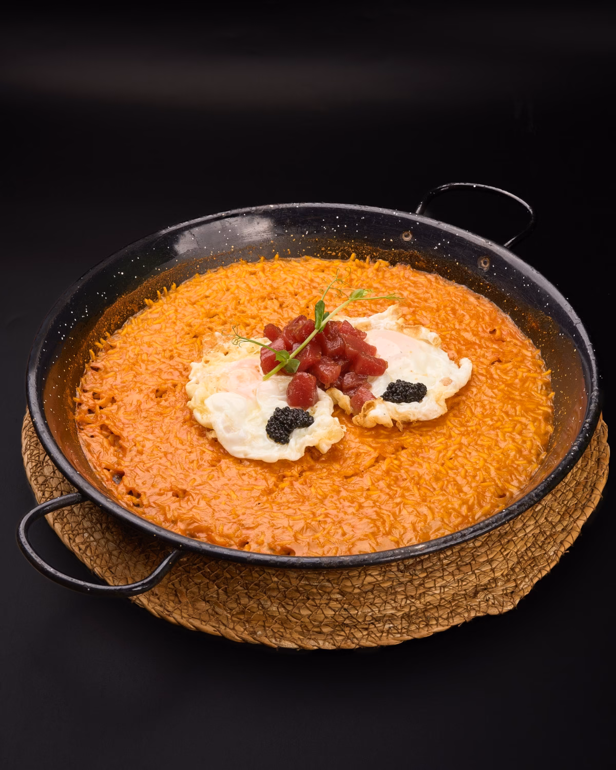 Pan of orange saffron risotto topped with two scoops of sour cream, red fruit pieces, and black caviar-like pearls, garnished with microgreens on a black skillet and woven mat.