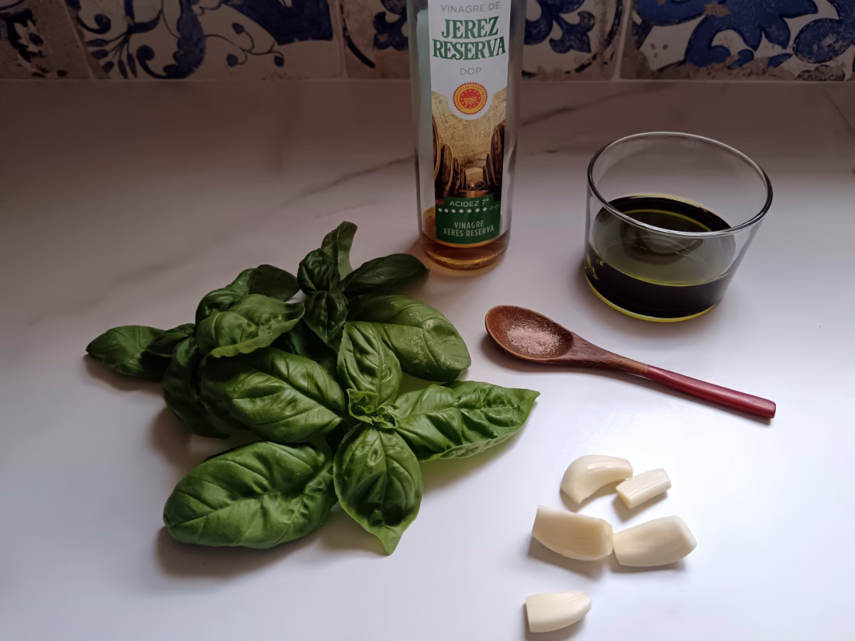 The ingredients needed to make basil garlic dressing on the countertop: basil, olive oil, garlic, salt, and sherry vinegar.