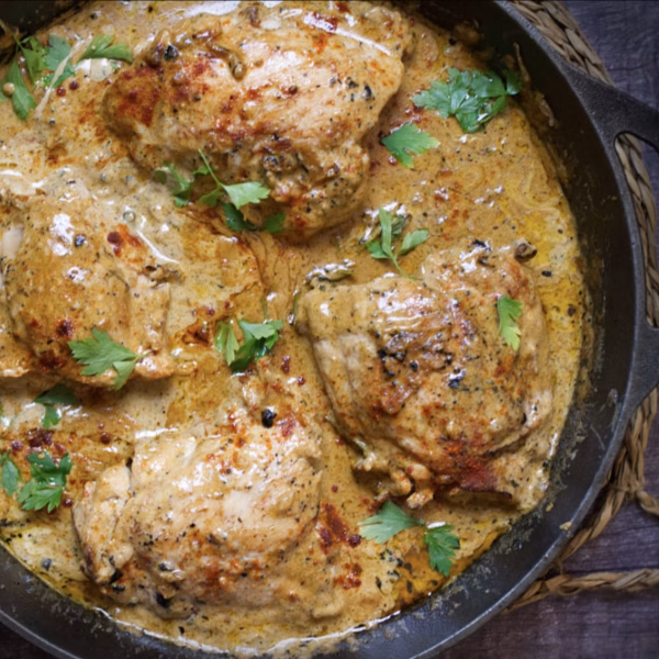 Four cooked chicken thighs in a skillet with creamy sauce and parsley flakes.