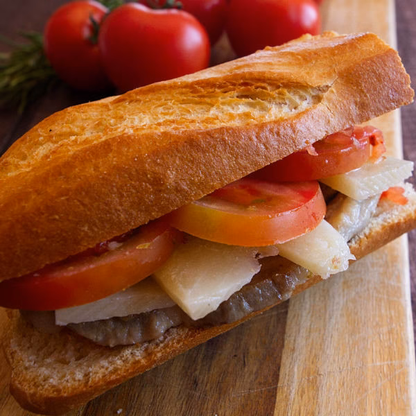 A sandwich on a chopping board with cooked pork loin, Manchego cheese, and slices of tomato.