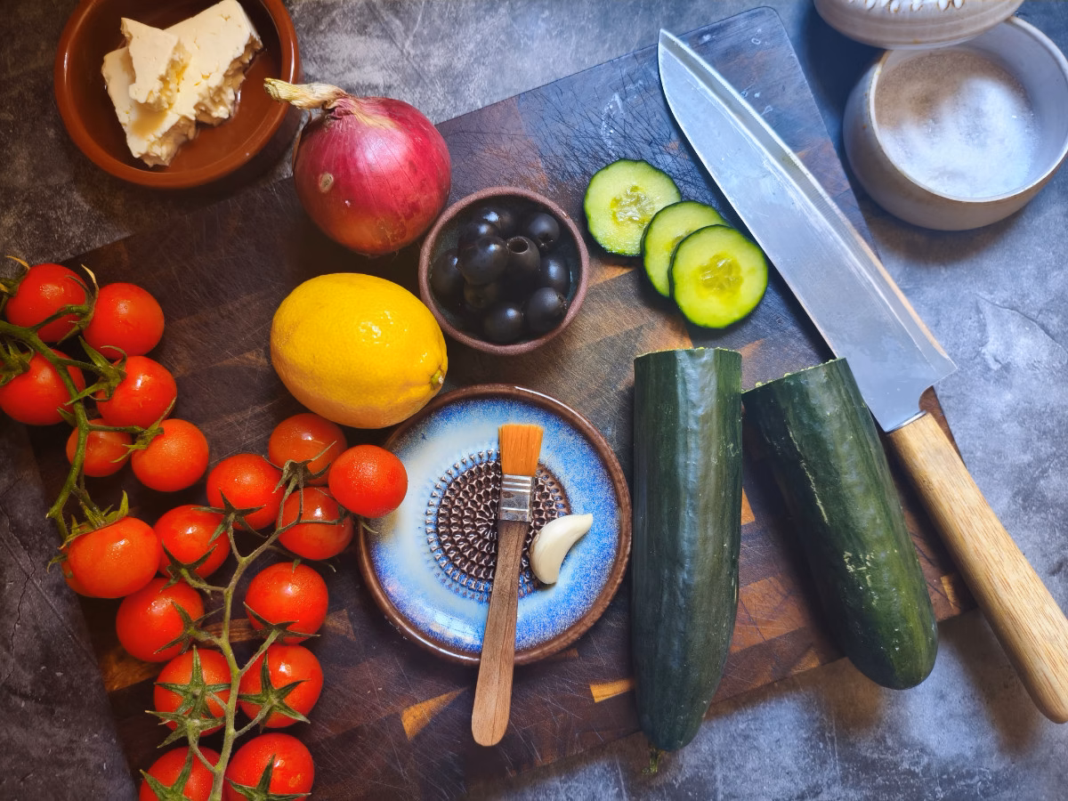 Ingredients used to make a Mediterranean cucumber salad are laid out on a wooden cutting board