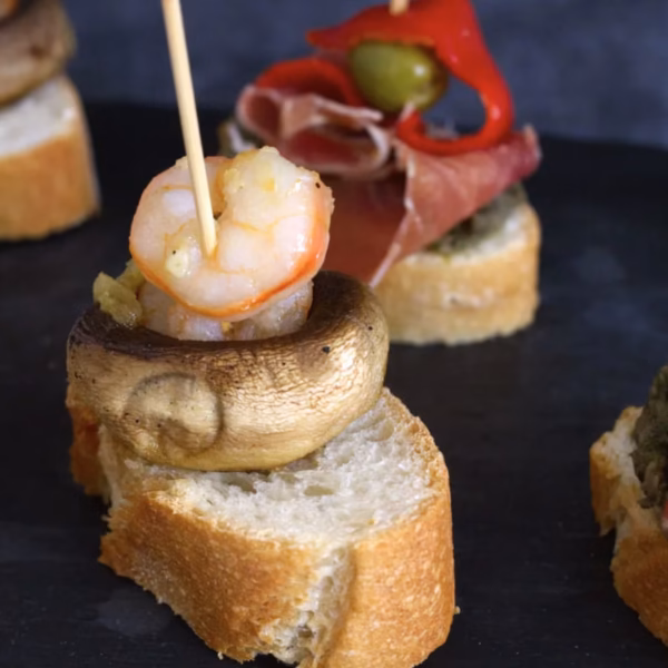 A pinchos with a prawn and mushroom and one in the background with ham, red pepper, and an olive.