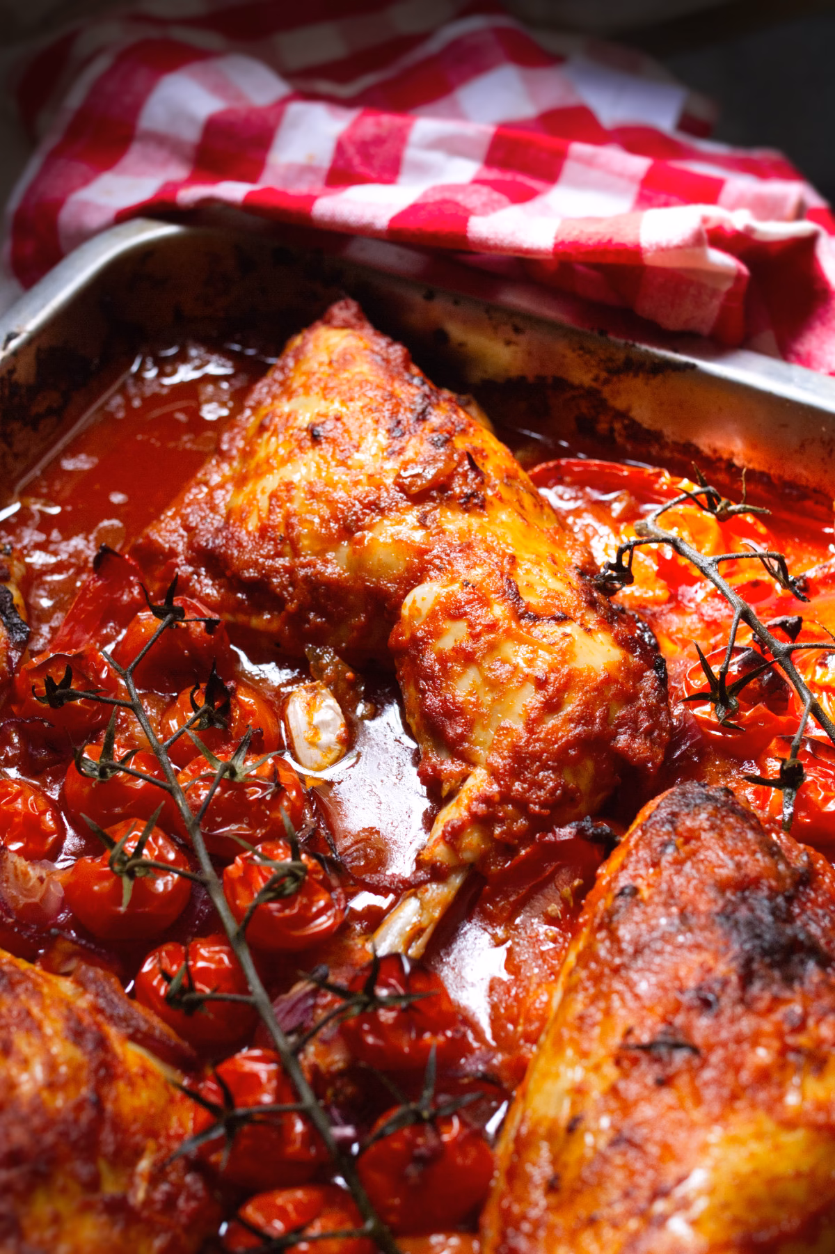Roasted chicken in a red tomato sauce with cherry tomatoes and thyme, in a metal pan with a red-and-white checkered cloth nearby