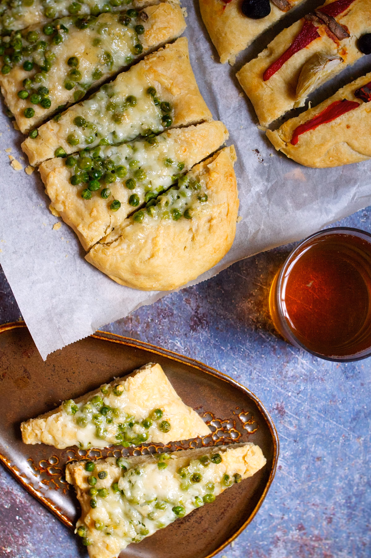 Spanish Cocas-v49 Savory cheese pastry squares topped with green peas on parchment paper. nearby a glass of red sauce.